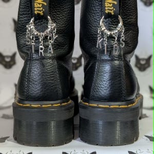 Könnte beinhalten: Schwarze Leder-Plateau-Stiefel mit gelben Nähten und einer schwarzen Sohle. Silberne Charms buchstabieren "STRA" und "KIDS" auf der Rückseite jedes Stiefels. Die Stiefel haben eine strukturierte Lederoberfläche und ein gelbes "Air Wair"-Etikett.
