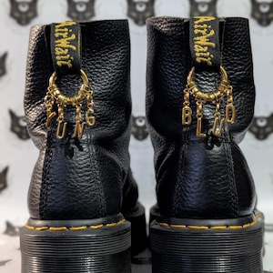 May include: Black leather platform boots with gold charms spelling "YUNG BLUD". The boots feature yellow stitching and a black sole. The boots have a textured leather upper and a branded pull tab.