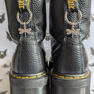 Puede incluir: Botas plataforma de cuero negro con costuras amarillas y un charm de libélula unido a los cordones. Las botas tienen una suela de plataforma gruesa y una lengüeta de tiro con el texto "Dr. Martens" en ella.