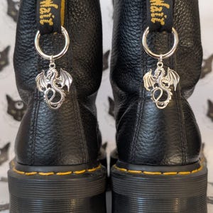Könnte beinhalten: Ein Paar schwarze Lederstiefeletten mit strukturierter Oberfläche und dicken Sohlen. Jeder Stiefel hat eine gelb-schwarze Fersenzuglasche mit der Aufschrift AirWair, einen silbernen Metallring und einen silbernen Drachenanhänger an der Ferse.