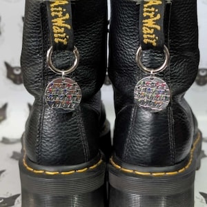 Op de afbeelding: Zwarte leren laarzen met gele stiksels en een dikke zool. Elke laars heeft een geel en zwarte stoffen lus met "Air Wair" en een zilveren bedel met kleurrijke letters.