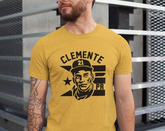 May include: A yellow t-shirt with a black graphic of baseball player Roberto Clemente wearing a cap with the number 21. The graphic also includes the text "CLEMENTE" and "PR".