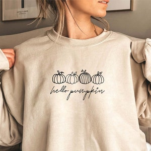 May include: Beige sweatshirt featuring a black line drawing of four pumpkins and the words "hello pumpkin". The top has long sleeves and is a neutral colour, suitable for autumn.