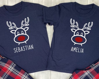 May include: Two navy blue short-sleeved shirts with a white reindeer graphic and the names "Sebastian" and "Amelia". Paired with red, white, and navy plaid pajama pants. The shirts have a red nose on the reindeer.