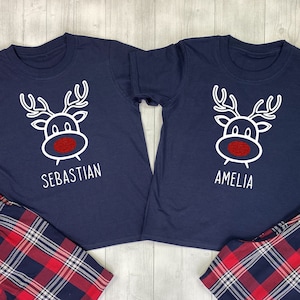 May include: Two navy blue short-sleeved shirts with a white reindeer graphic and the names "Sebastian" and "Amelia". Paired with red, white, and navy plaid pajama pants. The shirts have a red nose on the reindeer.