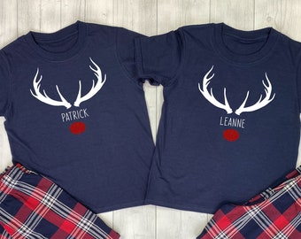 May include: Two navy blue short-sleeved t-shirts paired with red, navy, and white plaid pajama pants. Each shirt features a white graphic of reindeer antlers, a red glittery nose, and the printed names Patrick and Leanne respectively.
