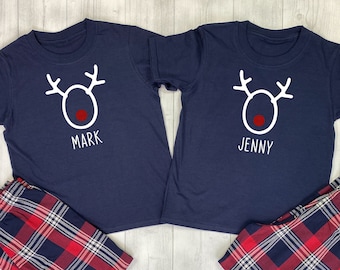 May include: Two navy blue t-shirts with a white reindeer design and the names "Mark" and "Jenny" printed below. The shirts are paired with red and black checked pyjama trousers.