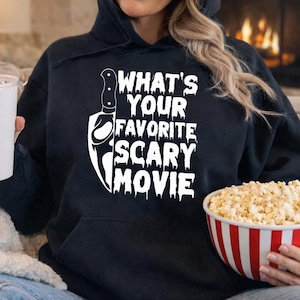 May include: Black hoodie with the text "WHAT'S YOUR FAVORITE SCARY MOVIE" and a knife graphic. A white cup with a red straw is held in one hand, and a red and white striped bowl of popcorn is held in the other.