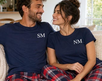 May include: Two navy blue short-sleeved t-shirts with white embroidered initials on the chest, paired with red, navy blue, and white plaid pajama pants. One shirt features the letters SM, while the other shirt features the letters NM.