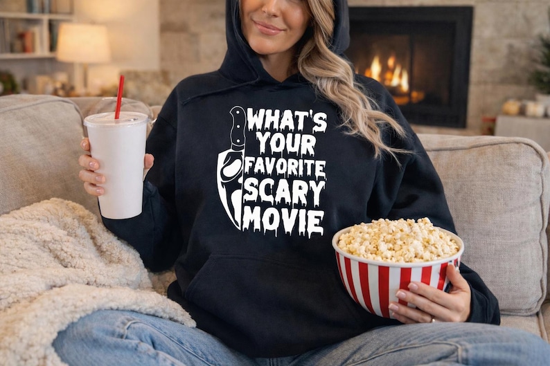 May include: Black hoodie with the text "WHAT'S YOUR FAVORITE SCARY MOVIE" and a knife graphic. A white cup with a red straw is held in one hand, and a red and white striped bowl of popcorn is held in the other.