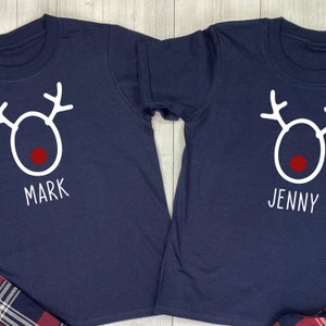 May include: Two navy blue t-shirts with a white reindeer design and red nose. The shirts have the names "Mark" and "Jenny" written below the reindeer.