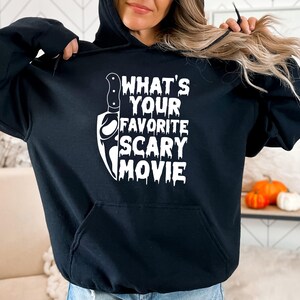 May include: Black hooded sweatshirt with a white graphic that says "WHAT'S YOUR FAVORITE SCARY MOVIE" and a knife illustration. The sweatshirt has a front pocket and a drawstring hood. The design is perfect for Halloween or horror movie fans.