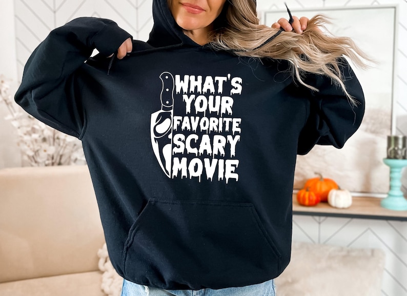 May include: Black hooded sweatshirt with a white graphic that says "WHAT'S YOUR FAVORITE SCARY MOVIE" and a knife illustration. The sweatshirt has a front pocket and a drawstring hood. The design is perfect for Halloween or horror movie fans.
