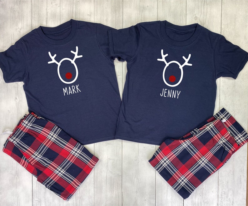May include: Two navy blue t-shirts with a white reindeer design and the names "Mark" and "Jenny" printed below. The shirts are paired with red and black plaid pajama pants.
