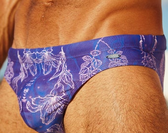 May include: Purple swim trunks with a white floral pattern. The trunks have a small logo on the side that says "Exotiq".