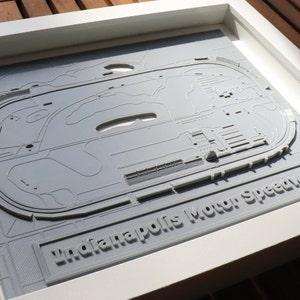 May include: A framed 3D printed model of the Indianapolis Motor Speedway race track. The model is gray and has a white frame. The text "Indianapolis Motor Speedway" is printed on the bottom of the frame.