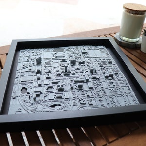 May include: A 3D printed city map in a black frame. The map is raised and shows the buildings and streets of a city. The map is on a wooden table.