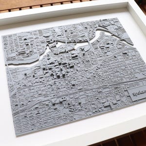May include: A framed 3D map of Spokane, Washington. The map is made of gray material and shows the city's streets and landmarks. The frame is white and has a simple design.