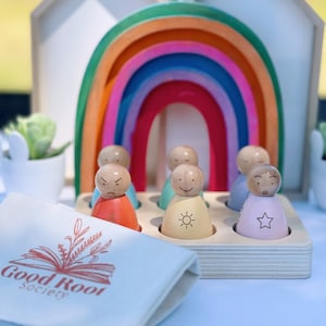May include: A set of wooden rainbow stacking arches with a rainbow-colored wooden archway.  The archway is decorated with small wooden figures with different facial expressions.  The figures are sitting in a wooden tray.  A white canvas bag with the text "Good Root Society" is in the foreground.