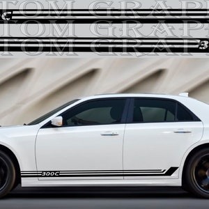May include: White Chrysler 300C sedan with black side stripes and the text "300C" on the side. The car is parked on a street with a gray wall in the background.