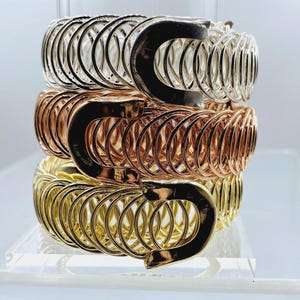 May include: A stack of three metallic bracelets, each featuring a series of interconnected circular wire loops and a smooth, curved, U-shaped clasp. The top bracelet is silver-toned, the middle is rose gold-toned, and the bottom is gold-toned.