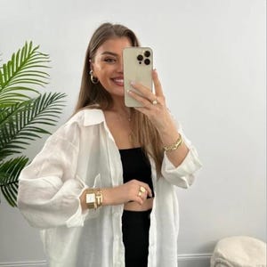 Könnte beinhalten: Weißes Leinenhemd mit langen Ärmeln, offen über einem schwarzen Top und einer schwarzen Hose getragen. Accessoires sind Goldschmuck, eine Uhr und ein Telefon.