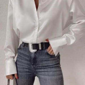 Könnte beinhalten: Ein weißes Satinhemd mit Kragen und langen Ärmeln, kombiniert mit einer blauen Jeans. Die Jeans wird mit einem schwarzen Gürtel mit silberner Schnalle getragen. Eine silberne Halskette ist ebenfalls zu sehen.