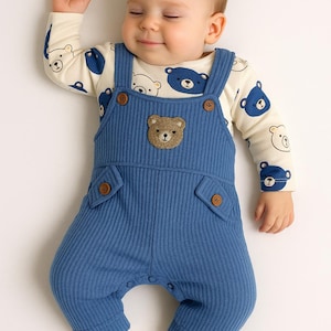 May include: A sleeping baby wearing a blue corduroy overall with a bear applique and a cream-colored long-sleeve shirt patterned with blue and white bears. The outfit has brown buttons and is paired with white socks.