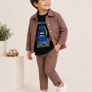 Könnte beinhalten: Ein Kind trägt ein mauvefarbenes Outfit, bestehend aus einer Jacke und Hose, kombiniert mit einem schwarzen T-Shirt mit einem Hai-Motiv und dem Wort "JAWS". Abgerundet wird das Ensemble durch weiße Turnschuhe, geeignet für den Alltag.