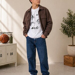 Könnte beinhalten: Eine Person trägt eine braune Jacke über einem weißen T-Shirt und einer blauen Jeans. Das T-Shirt hat den Text "It's not you, it's me" in schwarzer Schrift. Die Person trägt weiße Turnschuhe.