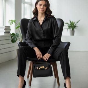 May include: A person in a black satin shirt and trousers, seated on a chair, holding a black handbag with a gold clasp. Black high heels are worn. The room is bright. The outfit is a classic, elegant style.