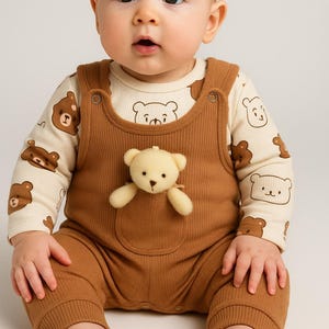 May include: A baby in a brown corduroy dungaree with a teddy bear in the pocket. The cream-coloured long-sleeved top has brown bear illustrations. The baby is looking at the camera.