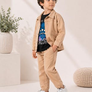 Könnte beinhalten: Beige Jacke und Hose, kombiniert mit einem schwarzen T-Shirt mit einem blauen Monster-Motiv und dem Text "AWS". Das Outfit wird durch weiße Turnschuhe ergänzt. Die Jacke hat eine Knopfleiste und die Hose hat Taschen.