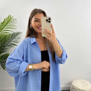 May include: A light blue button-down shirt with long sleeves and a chest pocket. The shirt is worn over a black top and paired with black pants. The person is wearing gold jewelry, including a watch, bracelets, earrings, and rings.