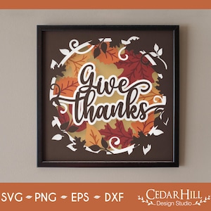 May include: A framed wall art piece with the words "Give thanks" in white script. The artwork features a circular design of fall leaves in shades of orange, red, and yellow, surrounded by white cut-out leaves and vines.