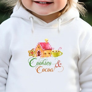 May include: A young child wearing a white hoodie and a matching knit hat with a pom-pom. The hoodie features a watercolor illustration of a gingerbread house, star cookie, and a cup of cocoa, with the words "Cookies & Cocoa" below.