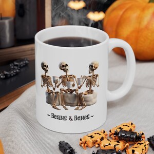 May include: White ceramic mug with a graphic of three skeletons sitting around a table drinking coffee. The mug has the text "Beans & Bones" printed on the bottom.