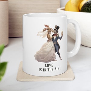 May include: White ceramic mug with a graphic of a rabbit and a cat dressed in wedding attire. The text "Love is in the air" is printed on the mug.