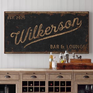 May include: A black and gold sign with the text "Wilkerson Bar & Lounge" and "Est. 2025". The sign has a distressed, vintage look and is hanging on a white wall.