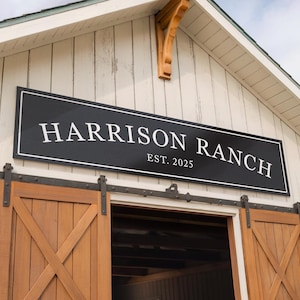 May include: A black rectangular sign with white lettering that reads "HARRISON RANCH EST. 2025" is mounted above a wooden barn door. The barn door is made of wood and has a cross-shaped design. The building is white with a wooden roof.