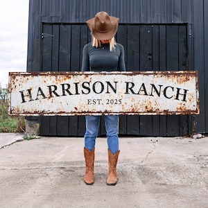 May include: A person holding a rusty metal sign that reads "HARRISON RANCH EST. 2025". The sign is white with black lettering.