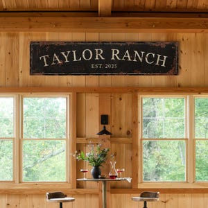 May include: A rustic wooden sign with black paint and white lettering reads "Taylor Ranch Est. 2025". The sign is hanging on a wooden wall with two windows behind it.