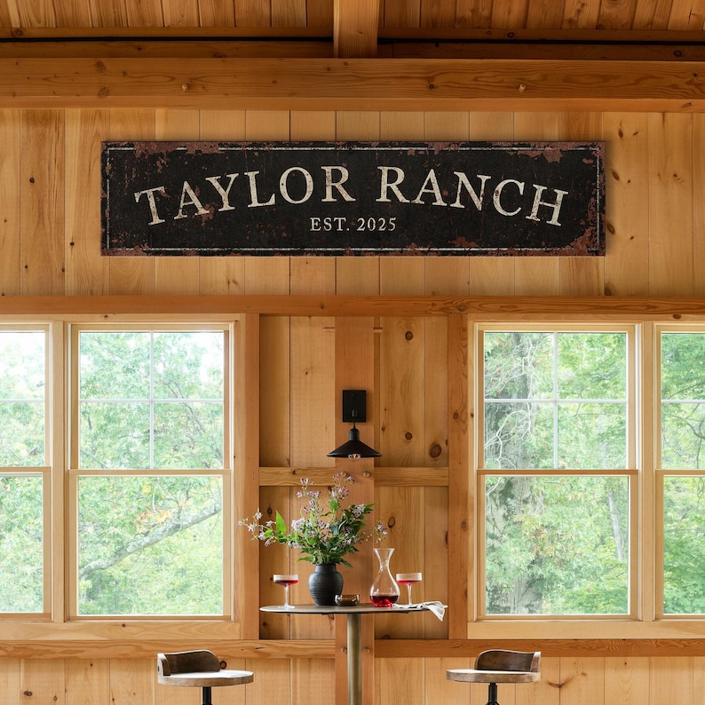 May include: A rustic wooden sign with black paint and white lettering reads "Taylor Ranch Est. 2025". The sign is hanging on a wooden wall with two windows behind it.