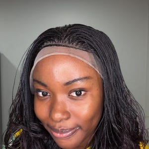 May include: A black lace front wig featuring micro-braided hair texture. The wig has a sheer, neutral-toned lace panel along the front hairline, transitioning into fine, thin braids that cover the scalp area and flow into loose, wavy hair ends.