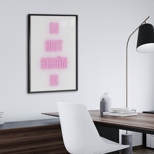 May include: A modern home office with a wooden desk, two white chairs, and a floor lamp. A framed print with the text "Du bist schon da" in pink neon lettering hangs on the wall.