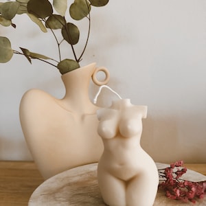 May include: A nude-colored candle shaped like a human torso. The candle is sitting on a wooden surface with a vase and dried flowers in the background.