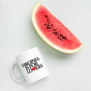 May include: A white coffee mug with the text "VIRGINIA IS FOR LOVERS" in black, with a red heart replacing the word "LOVE". A slice of watermelon with black seeds is next to the mug.