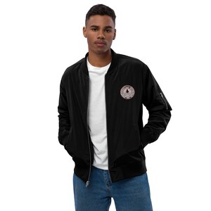 May include: A black bomber jacket with a zipper closure and a red and white embroidered patch on the left chest. The patch features a tree and the text "Adventure Awaits".