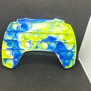 May include: A blue and green pop it fidget toy shaped like a video game controller. The toy has a white background with blue and green swirls.