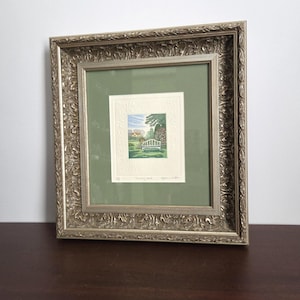 May include: Framed artwork featuring a serene garden scene with a white bench, trees, and a house. The artwork is set against a green background within a decorative silver frame. The frame has an ornate, textured design.